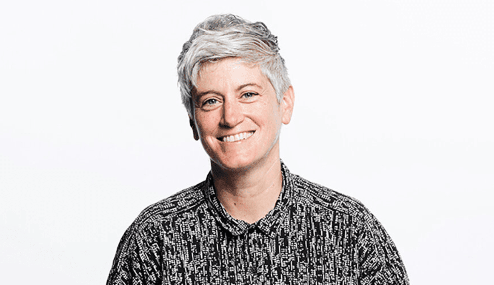 Individual with short, gray hair wearing a black and white patterned shirt against a plain background.