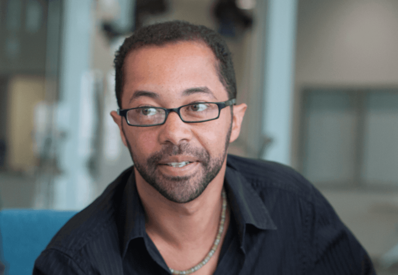 Man with glasses wearing a dark shirt looking to the side in a modern indoor setting.